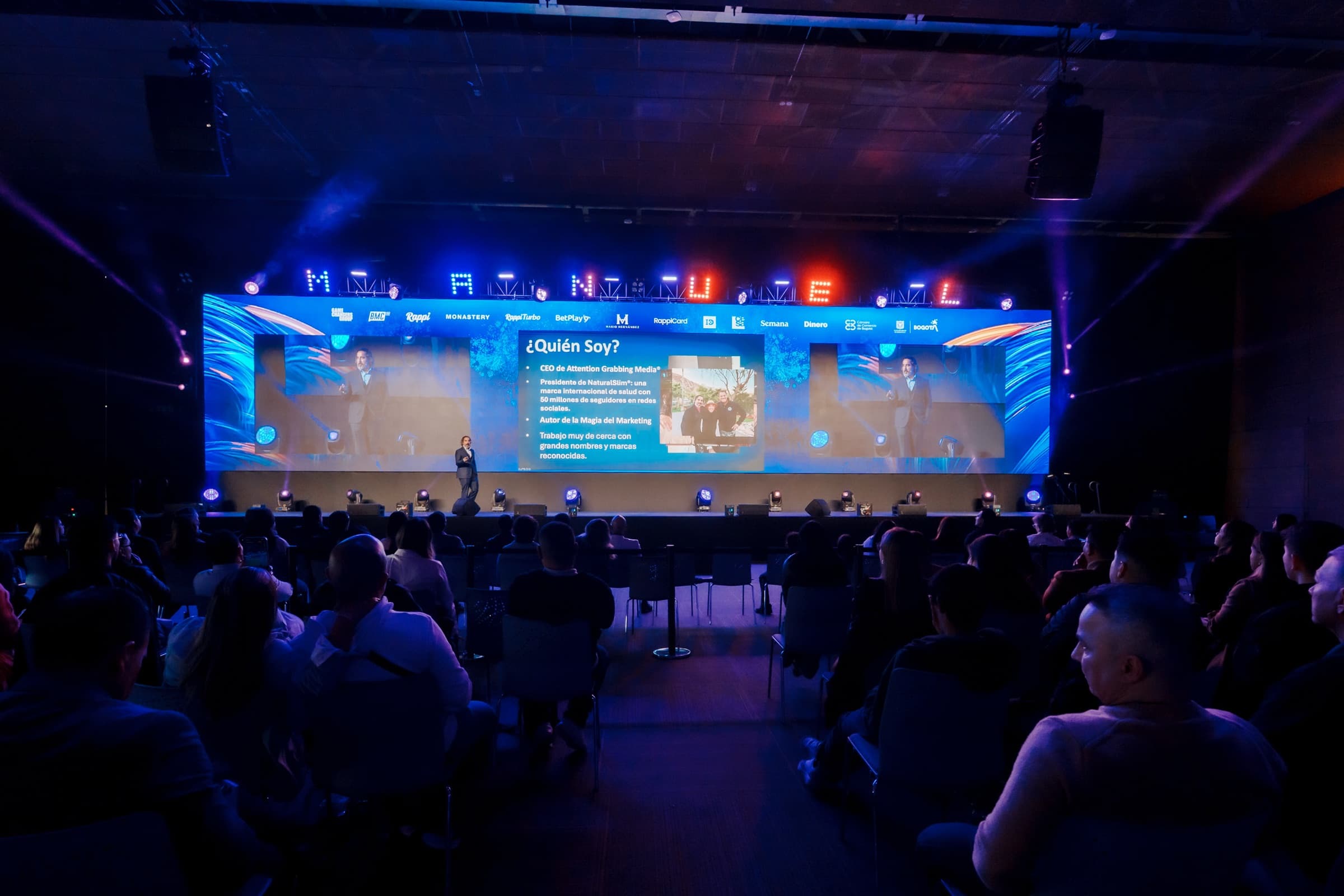 A wide view of a darkened Bogotá auditorium at Game Changers 2025, the full seated audience seen from the back rows while Manuel Suarez presents from a lit stage below a large LED wall.